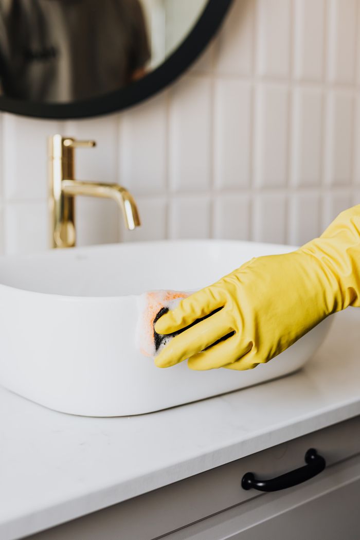 who-we-are Crop faceless person in yellow rubber protective glove wiping surface of white sink in bathroom with sponge and cleaning agent